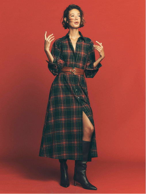 A model stands against a solid red studio background wearing a long-sleeved plaid shirt dress in black and red, styled with a brown belt and black knee-high boots.