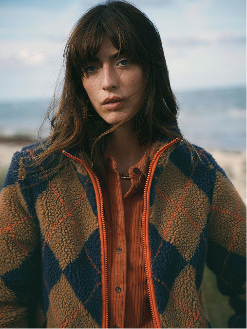 A woman wearing a brown and navy argyle sherpa fleece zip-up jacket over an orange button-up shirt, photographed outdoors with a blurred beach and sky background.
