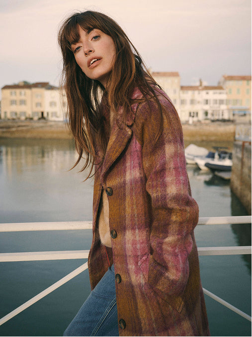 A woman wearing a brown and pink plaid coat with blue jeans, standing on a boat deck with a harbor and buildings in the background.