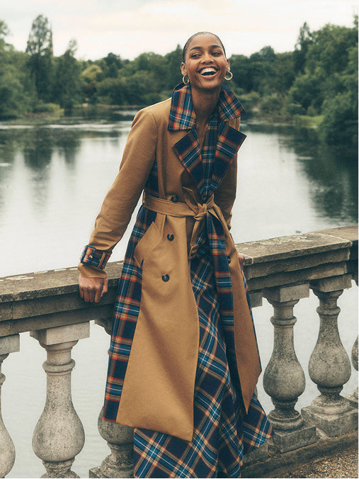 A woman stands on a stone bridge beside a lake, wearing a long tan trench coat with a plaid patterned lining and matching plaid collar, belt, and skirt. Trees and water are visible in the background.
