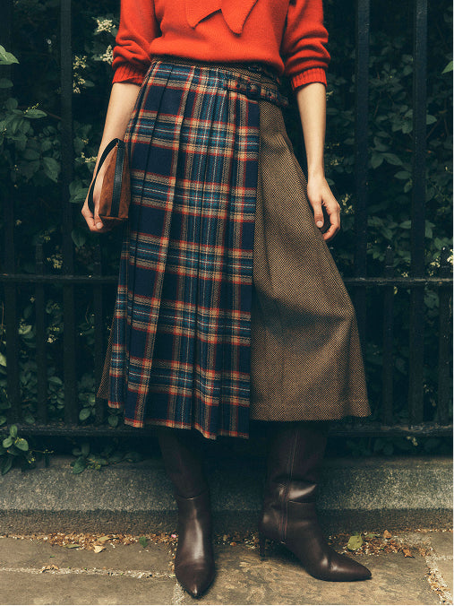 A person wearing a long pleated skirt with a split design: one half is navy, red, and orange plaid, and the other half is solid taupe-brown. The outfit is styled with a bright orange-red sweater, dark brown pointed-toe boots, and a brown handbag, standing outdoors in front of a black metal fence and green foliage.