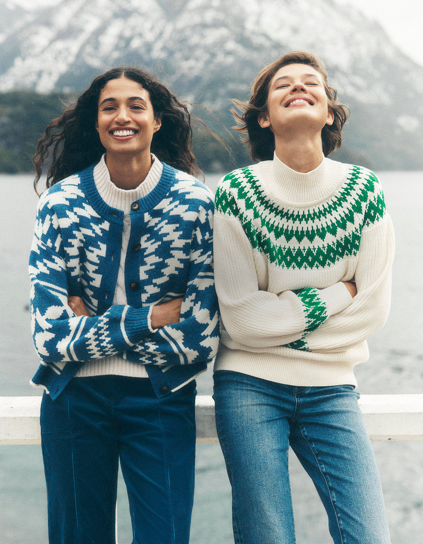 Two women wearing patterned sweaters standing by a body of water with mountains in the background.