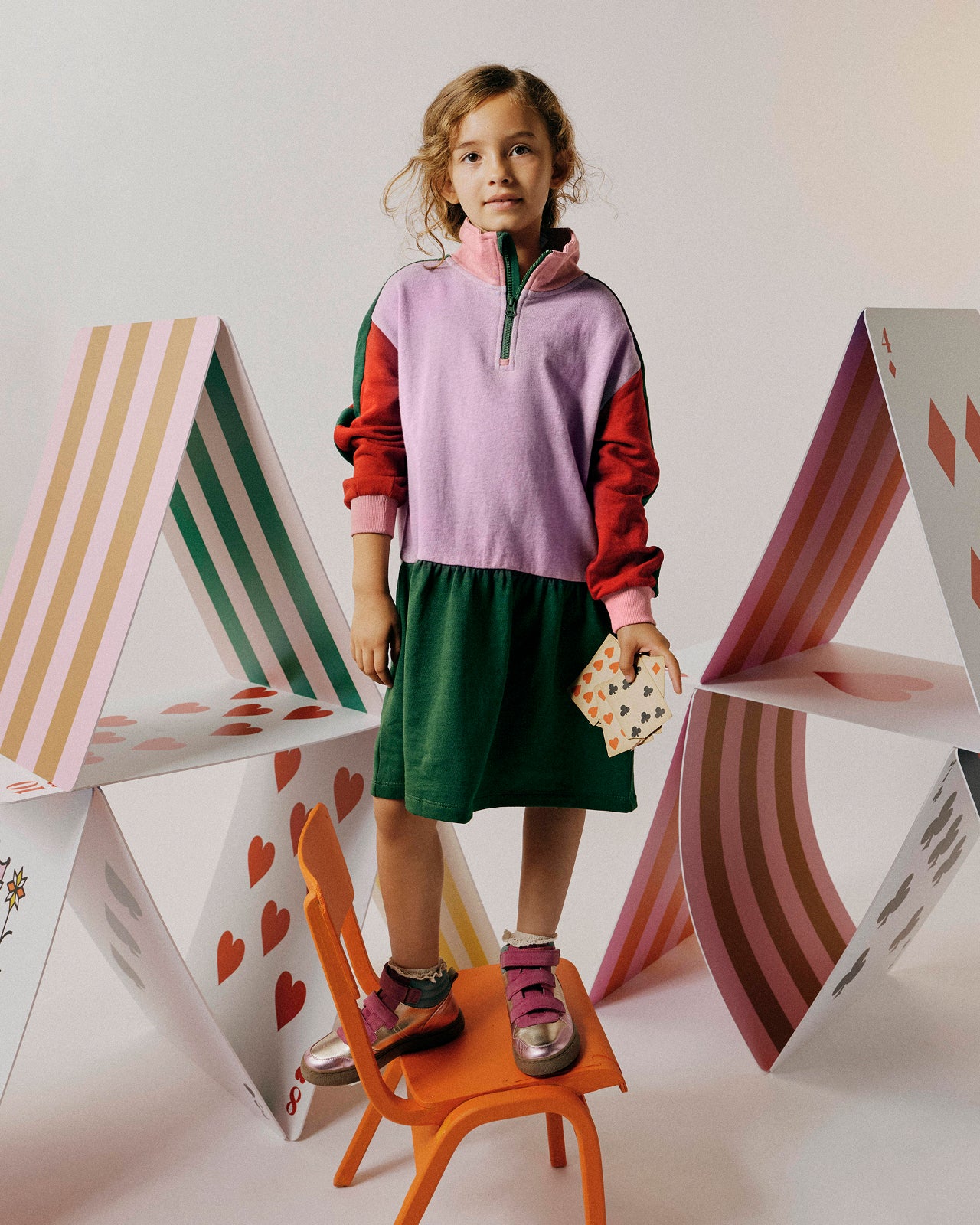 Child wearing a colorful outfit standing next to a geometric house-shaped chair.