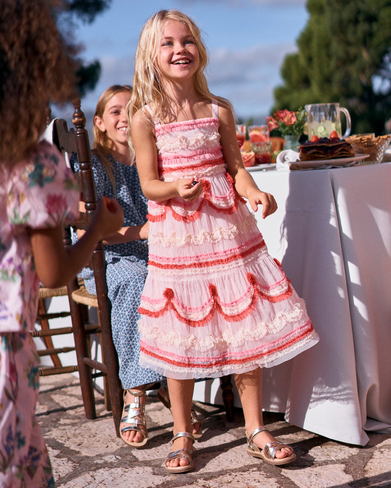 Young girl in a pink and white dress standing outdoors with people and tables in the background.