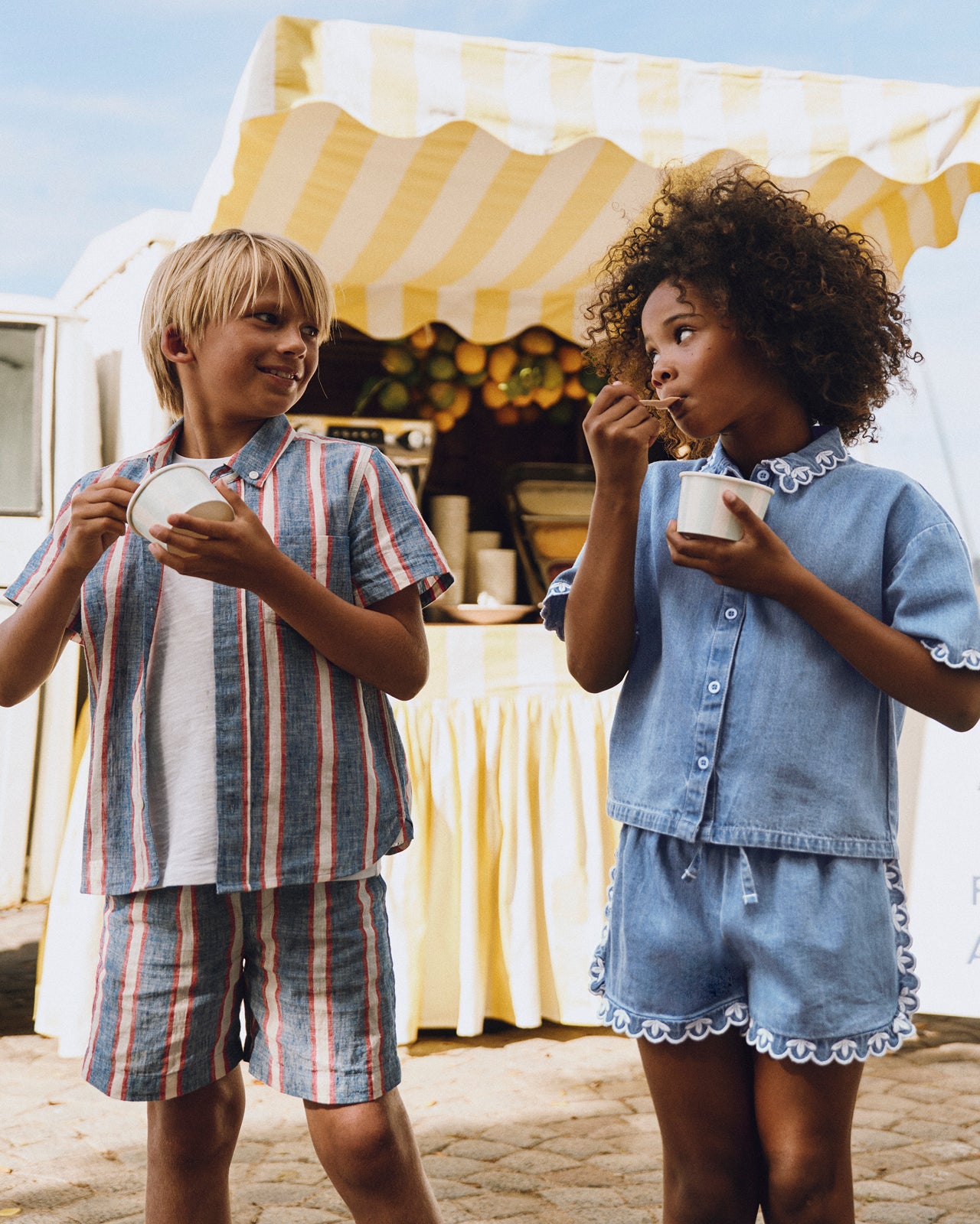 Two children in matching outfits eating from bowls outdoors with a striped canopy in the background.