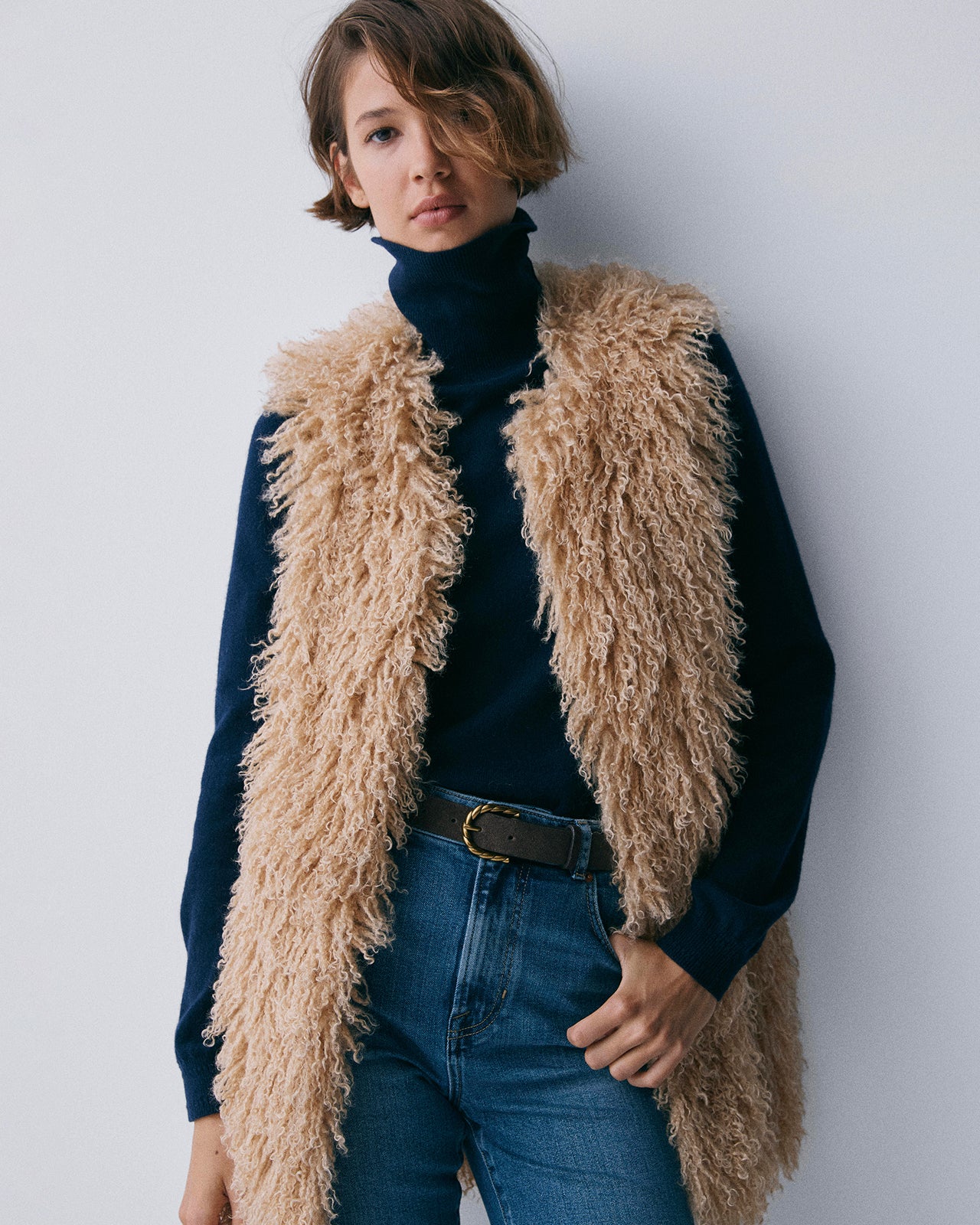 Person wearing a fluffy beige vest over a dark turtleneck and jeans against a white background