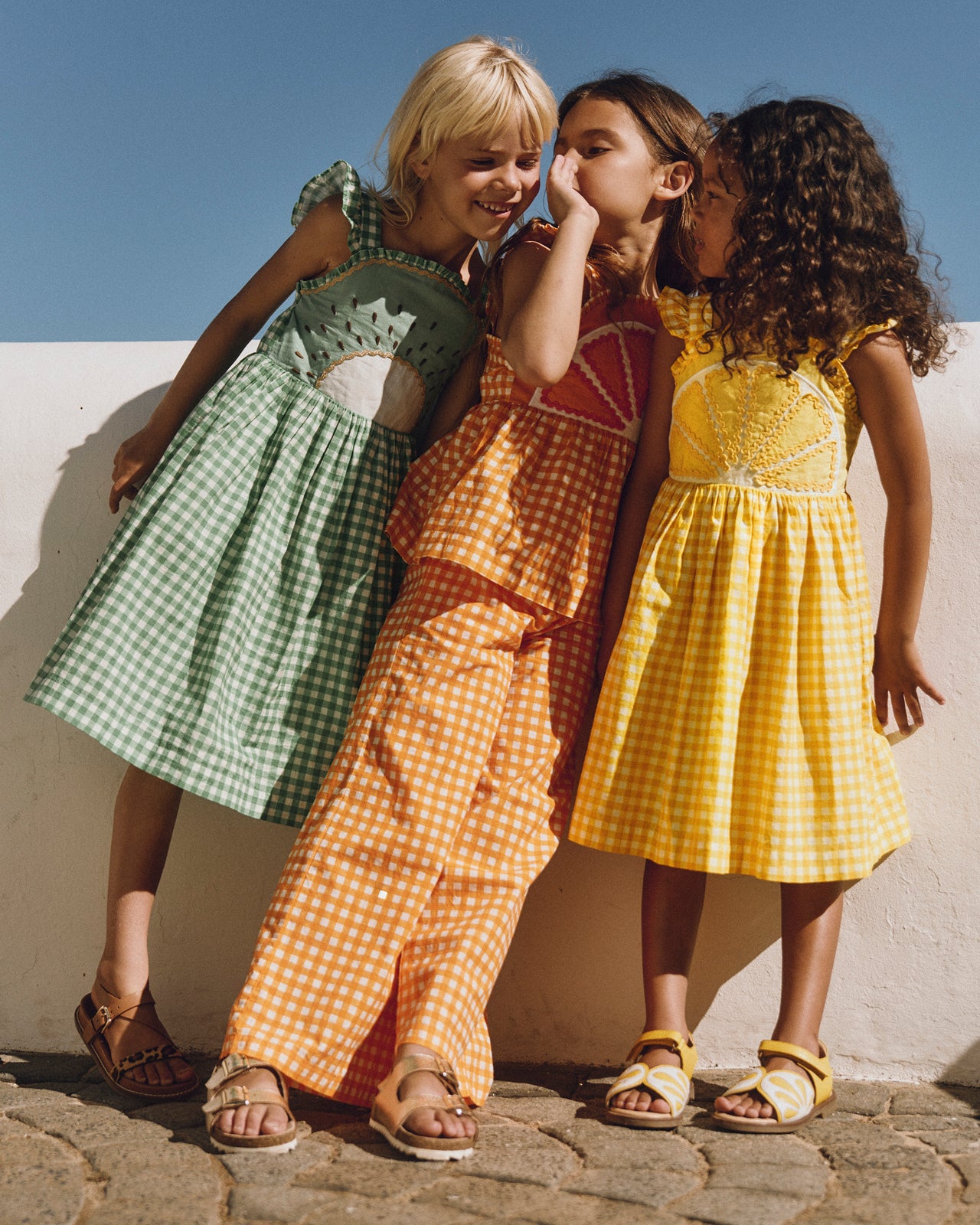 Three children in colorful dresses standing against a wall with a clear blue sky.