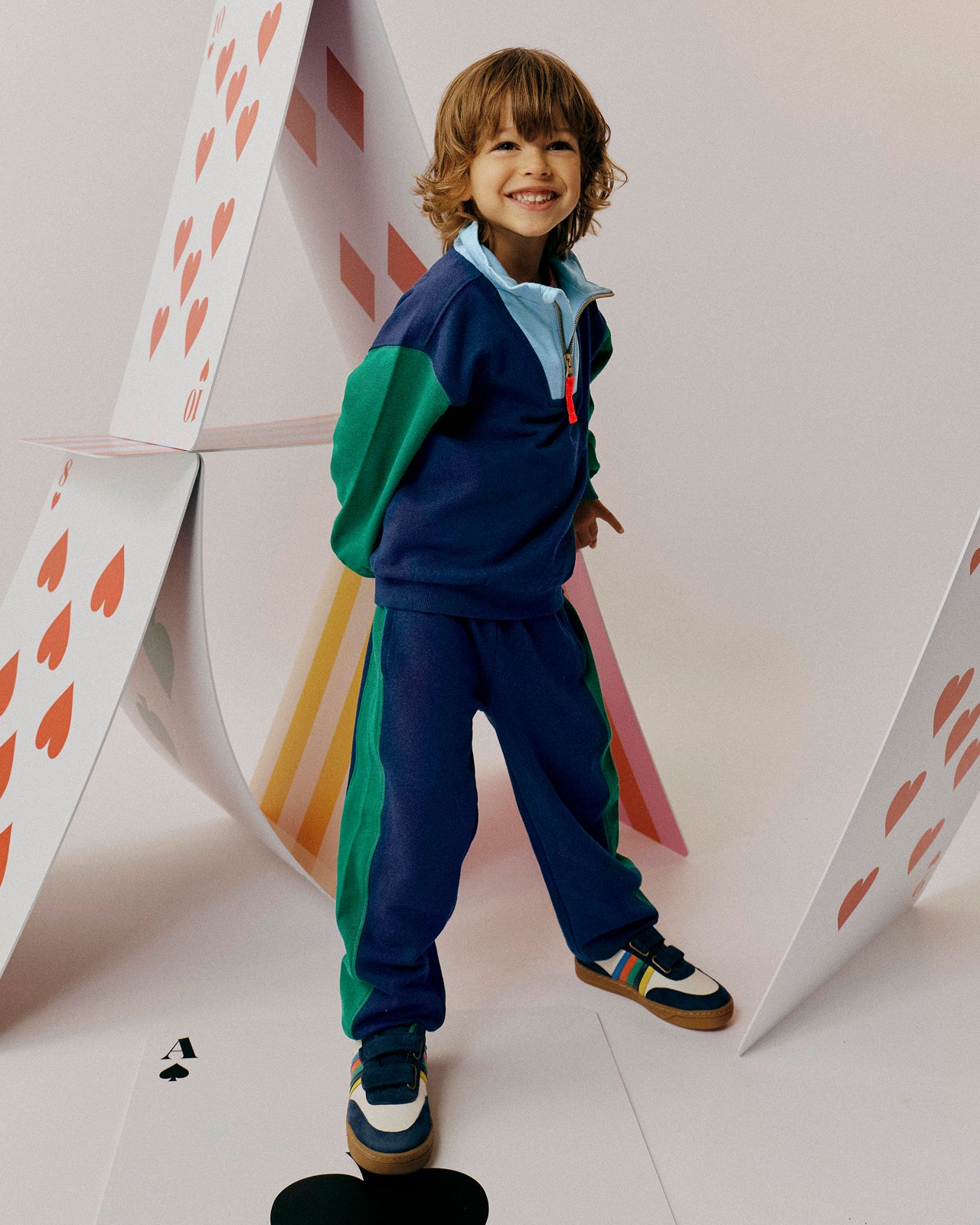 Child in a colorful outfit standing next to a large playing card with hearts on a white background