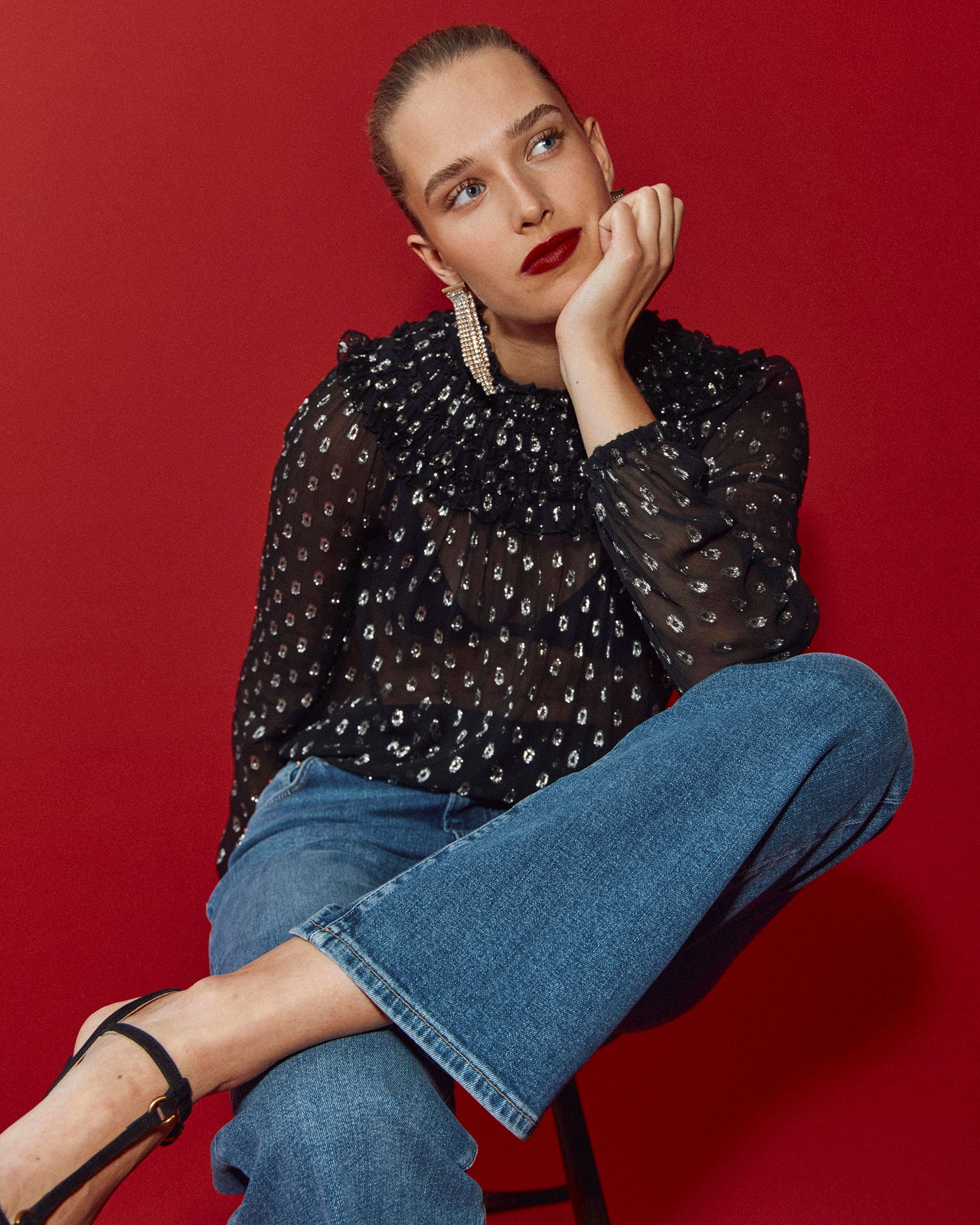Woman wearing a black sequin top and blue jeans sitting on a red background