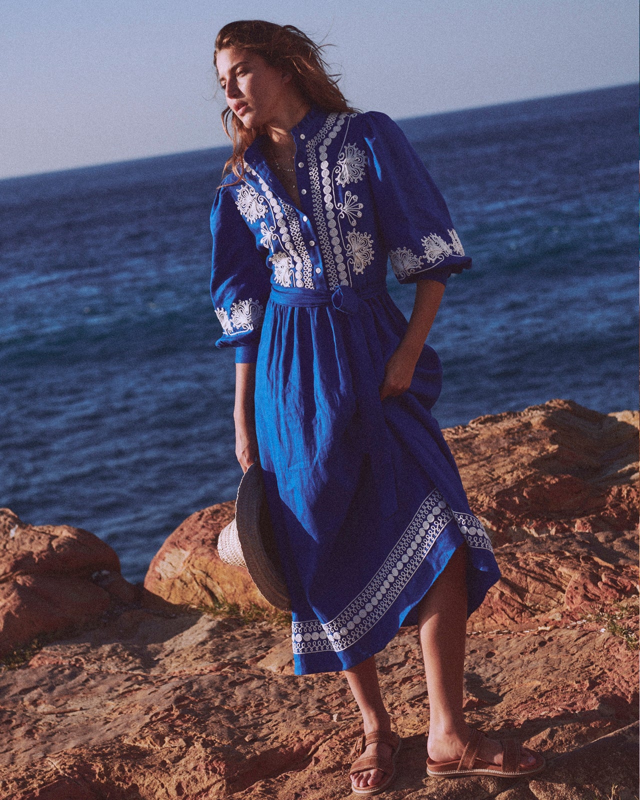 Woman in a blue dress with white embroidery standing on a rocky cliff overlooking the ocean.