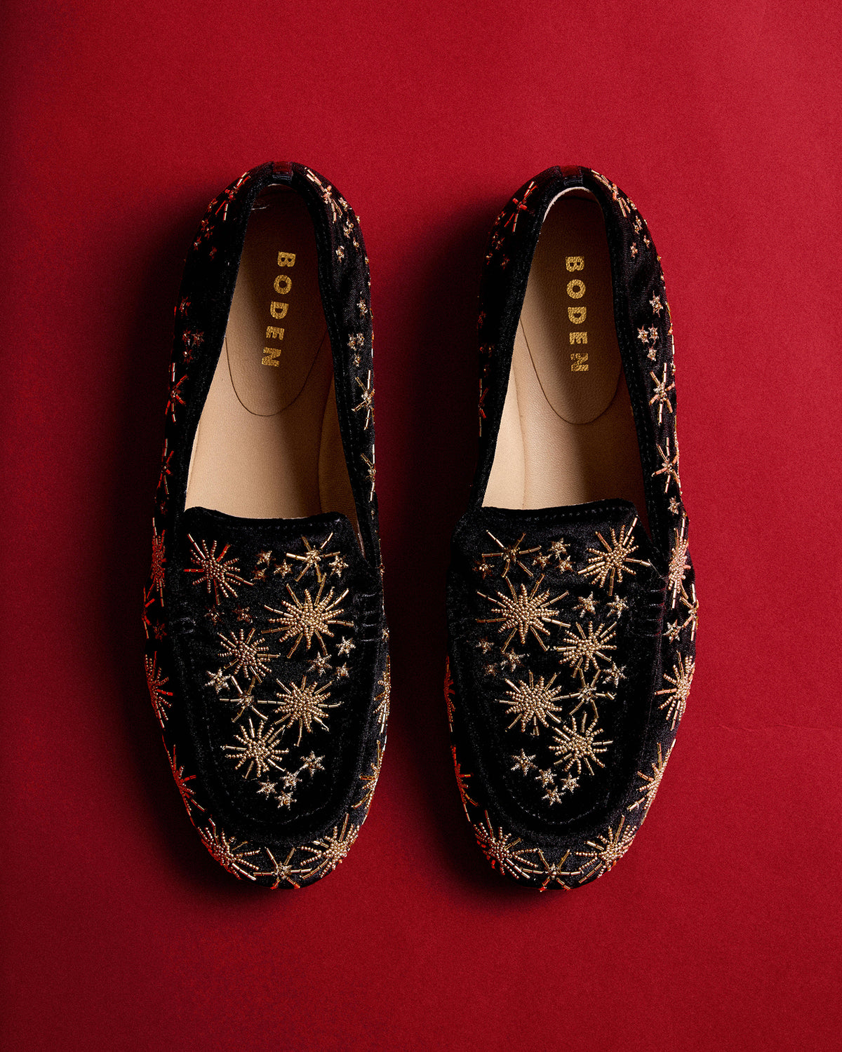 Pair of black velvet loafers decorated with intricate gold starburst and floral beadwork, photographed from above on a rich red background. Gold “BODEN” branding is visible inside each shoe.