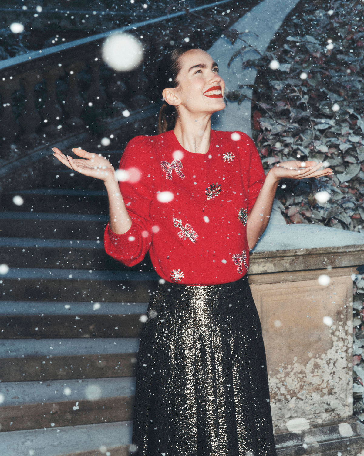 Women standing outdoors in falling snow, wearing a bright red jumper embellished with sparkling bows and festive motifs, paired with a shimmering black midi skirt. Stone steps and leafy foliage form the background.