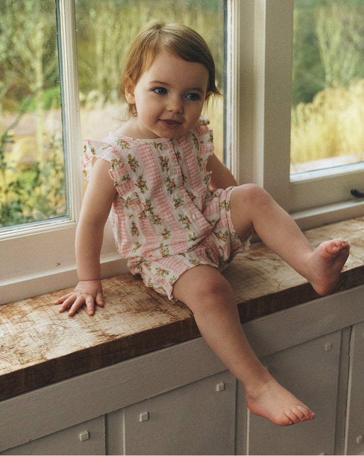 Child in a floral romper sitting on a window sill with a view of greenery outside.