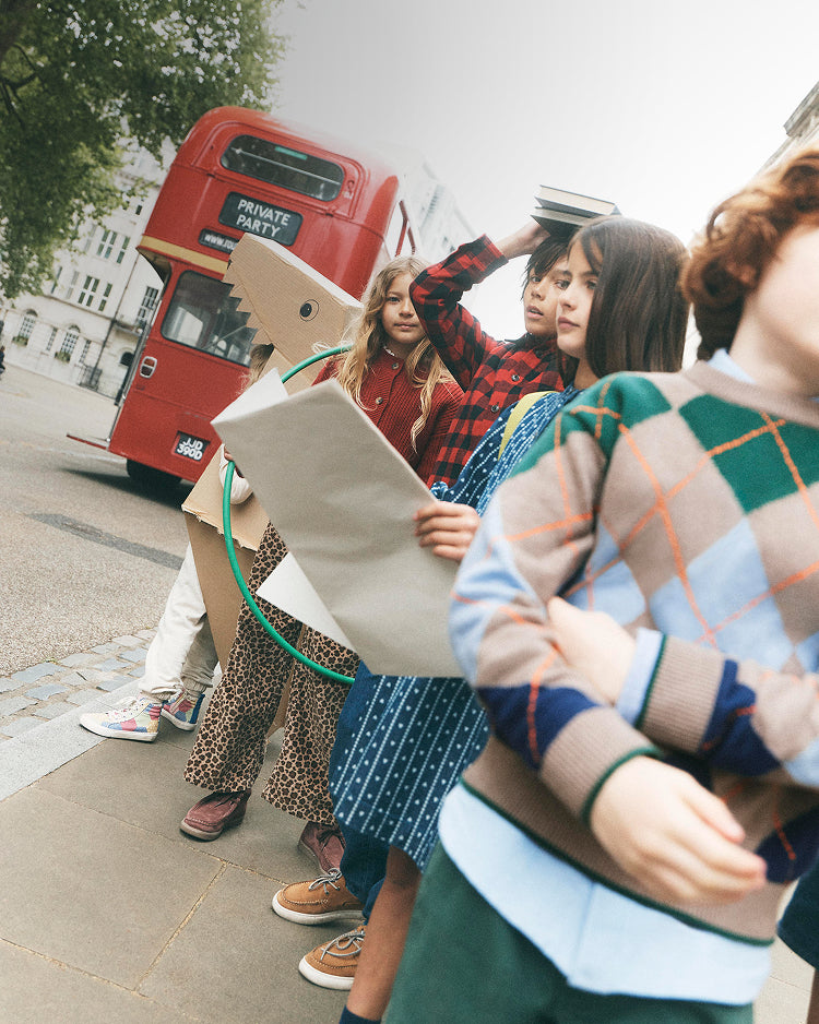 A group of children standing on a city sidewalk in front of a red double-decker bus. One child holds a large cardboard dinosaur head cutout, while another holds a green hula hoop.