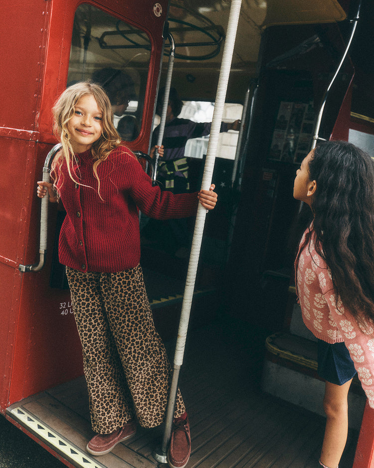 A young girl stands on the open door of a red bus, wearing a red knit cardigan and leopard-print pants, holding a grab handle. Another girl is visible in the doorway of the bus.