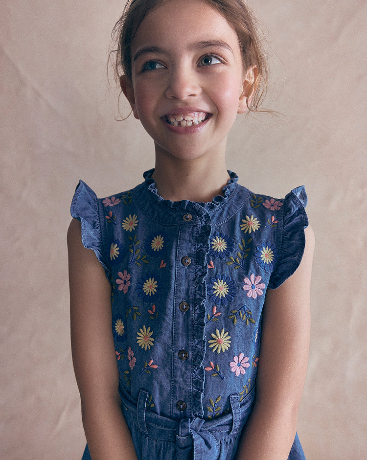 A young girl smiling and looking to the side, wearing a blue denim-style top with colorful embroidered flowers and ruffled sleeves, set against a plain beige backdrop.