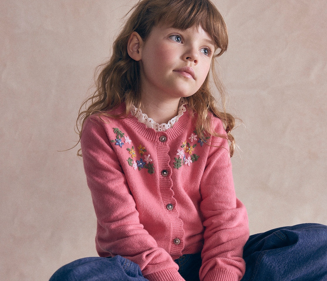 A young girl with light brown hair wearing a pink button-up cardigan with colorful floral embroidery and a ruffled white collar, paired with dark blue pants, sitting against a textured beige background.