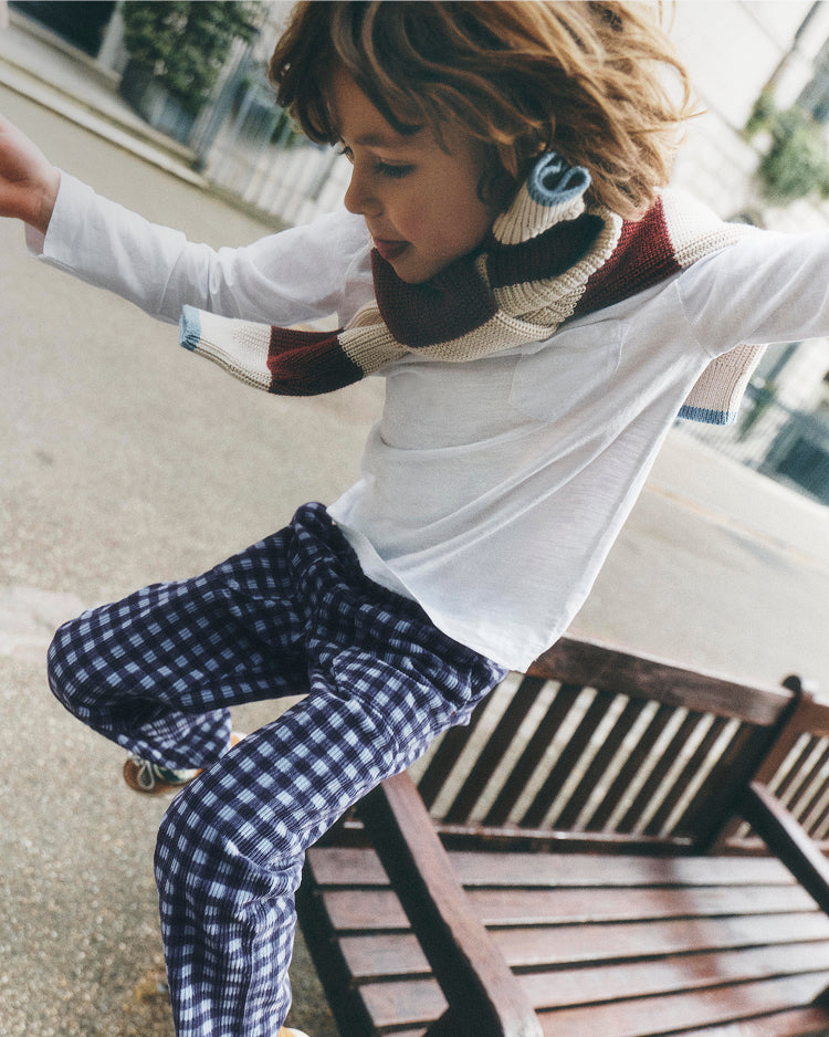 A child wearing a white long-sleeve shirt, a cream and burgundy knit scarf, and blue-and-white gingham pants, sitting on a wooden bench outdoors.