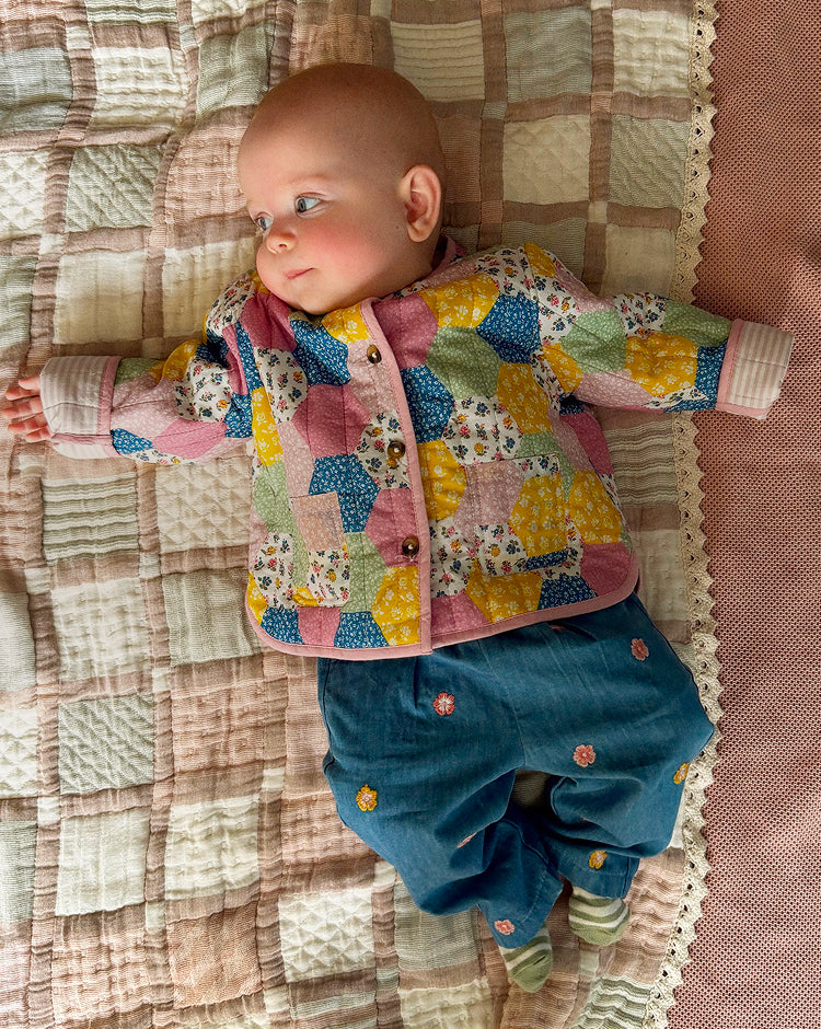 A baby lying on a quilted blanket, wearing a colorful patchwork-patterned cardigan and blue pants.