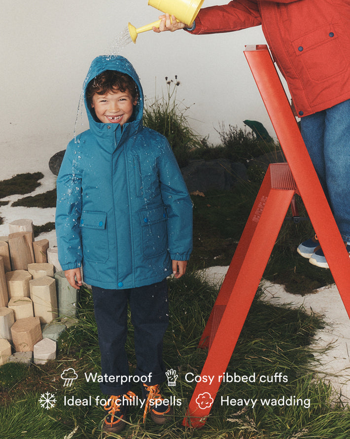 A child wearing a bright teal waterproof hooded jacket stands on grass while water is poured over the shoulders from above by an adult in a red jacket on a red step ladder. Product text overlays at the bottom mention waterproof, cosy ribbed cuffs, ideal for chilly spells, and heavy wading.