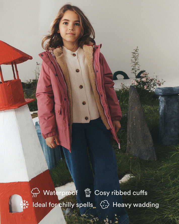 A young girl wearing a dusty rose pink hooded puffer jacket with a beige sherpa lining, a cream ribbed knit cardigan, and dark blue jeans, standing in a styled outdoor play area with a lighthouse prop. Text overlays at the bottom highlight product features like waterproofing and cozy cuffs.