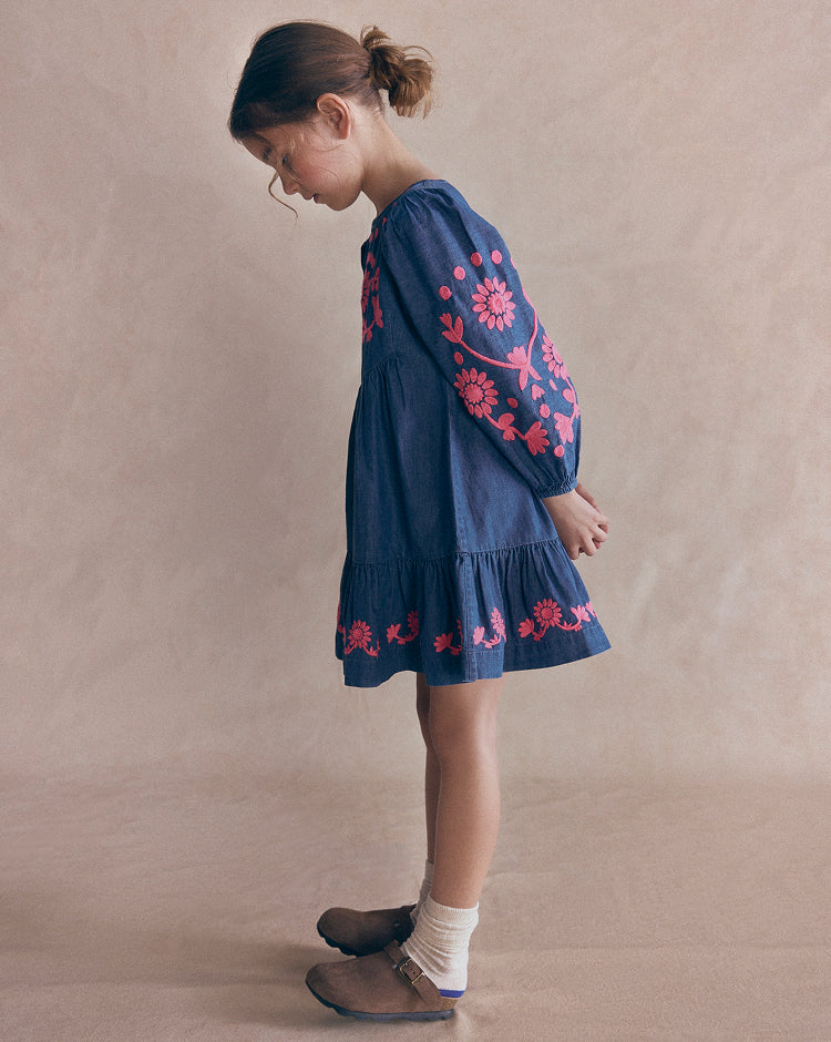 A young girl wearing a dark blue denim dress with pink floral embroidery, standing in profile against a neutral beige studio backdrop.