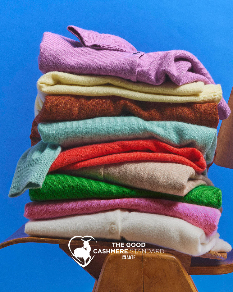 Stack of colorful cashmere sweaters on a wooden stool with a blue background, featuring 'The Good Cashmere Standard' brand.