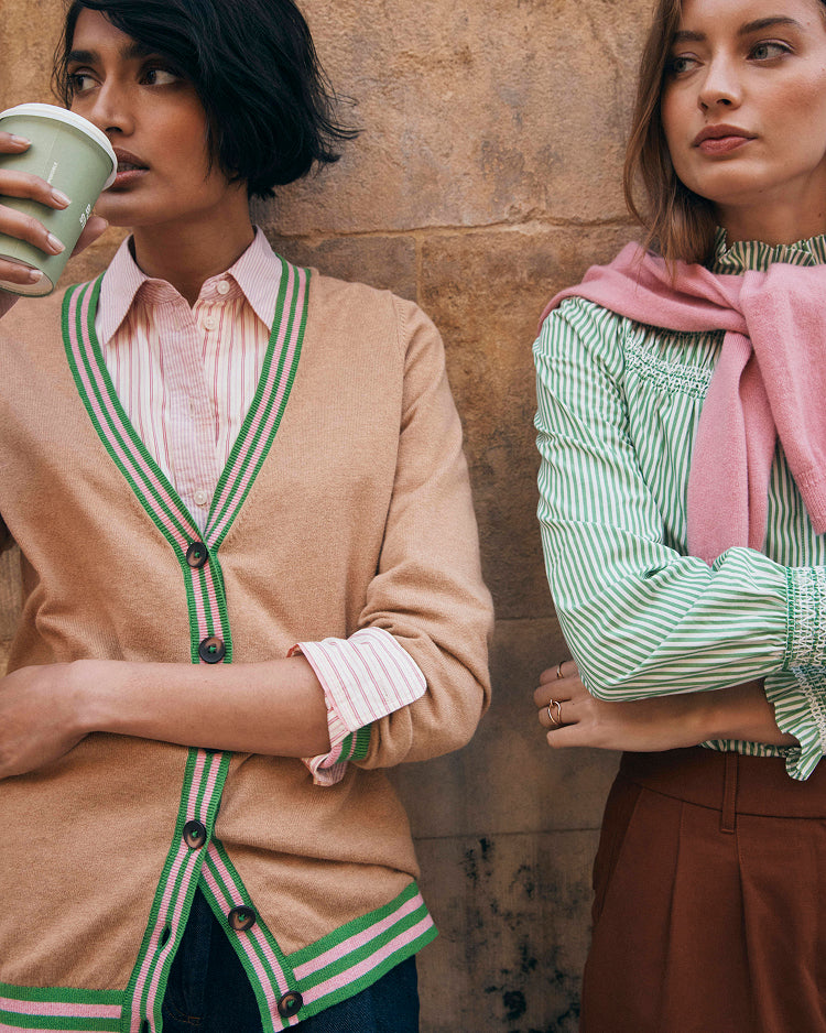 Two women standing against a stone wall, one holding a cup, both wearing stylish outfits.
