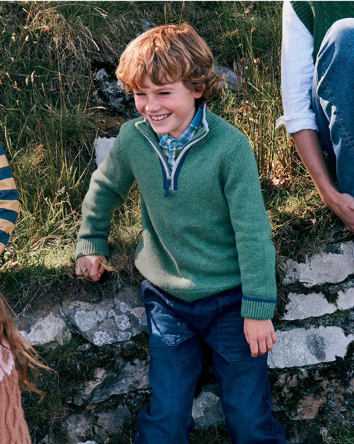 Child in a green sweater and blue pants standing on a stone path with grass and rocks in the background.