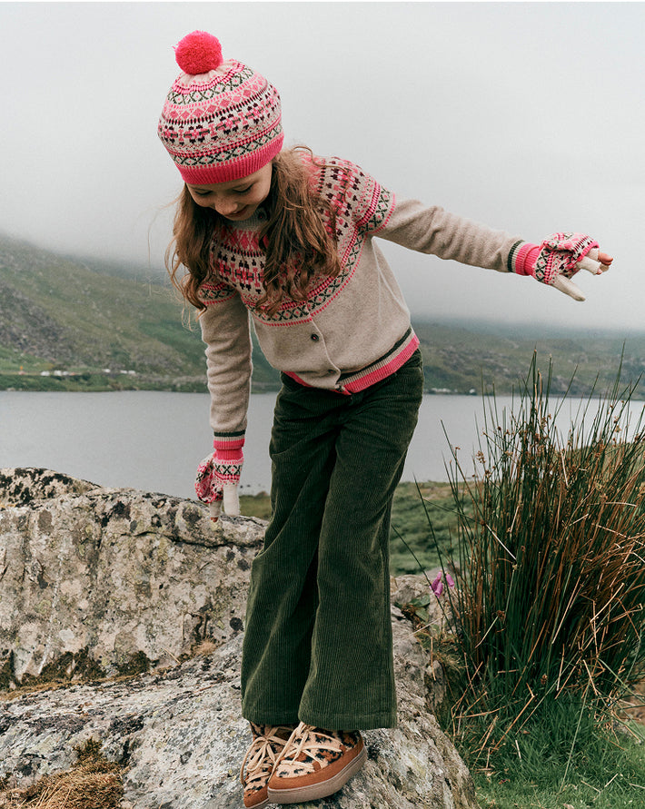 Person wearing a patterned sweater and pink hat with a pom-pom, standing on rocks by a lake.