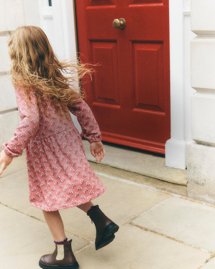 A young girl with long blonde hair is seen from behind, wearing a pink patterned long-sleeve dress and dark brown Chelsea boots, walking towards a red door on a stone sidewalk.