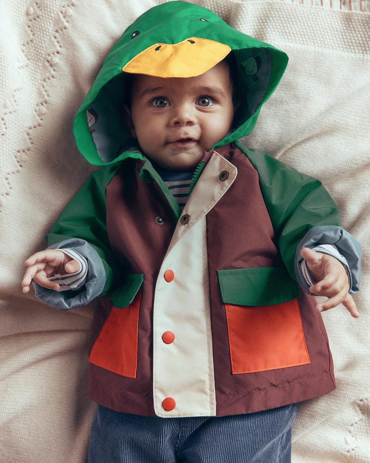 A baby wearing a colorful duck-themed hooded raincoat, lying on a textured cream blanket. The raincoat features a green hood with a yellow duck face, green sleeves, a brown body, a cream front panel, and orange pocket details.
