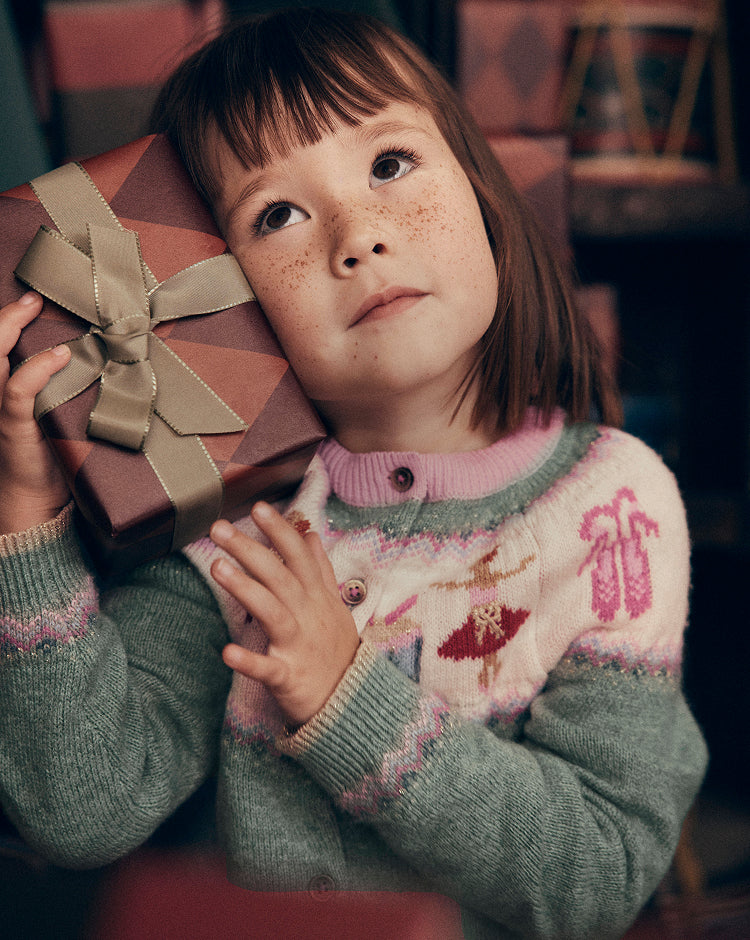 Close-up of a child holding a wrapped gift with a gold ribbon, wearing a Fair Isle knit cardigan in soft pink, cream and sage green with festive motifs including ballet shoes and dancers. The image highlights the detailed knit pattern and cosy texture against a warm indoor background.