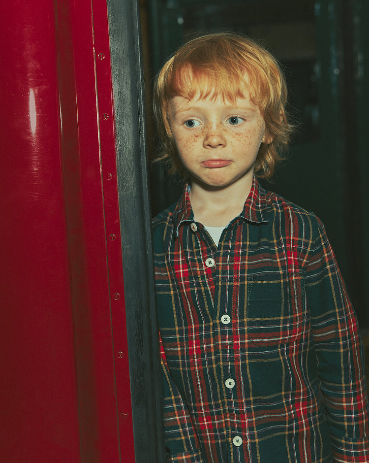 A young boy with red hair and freckles wearing a red, green, and black plaid button-up shirt, standing next to a red metal structure.
