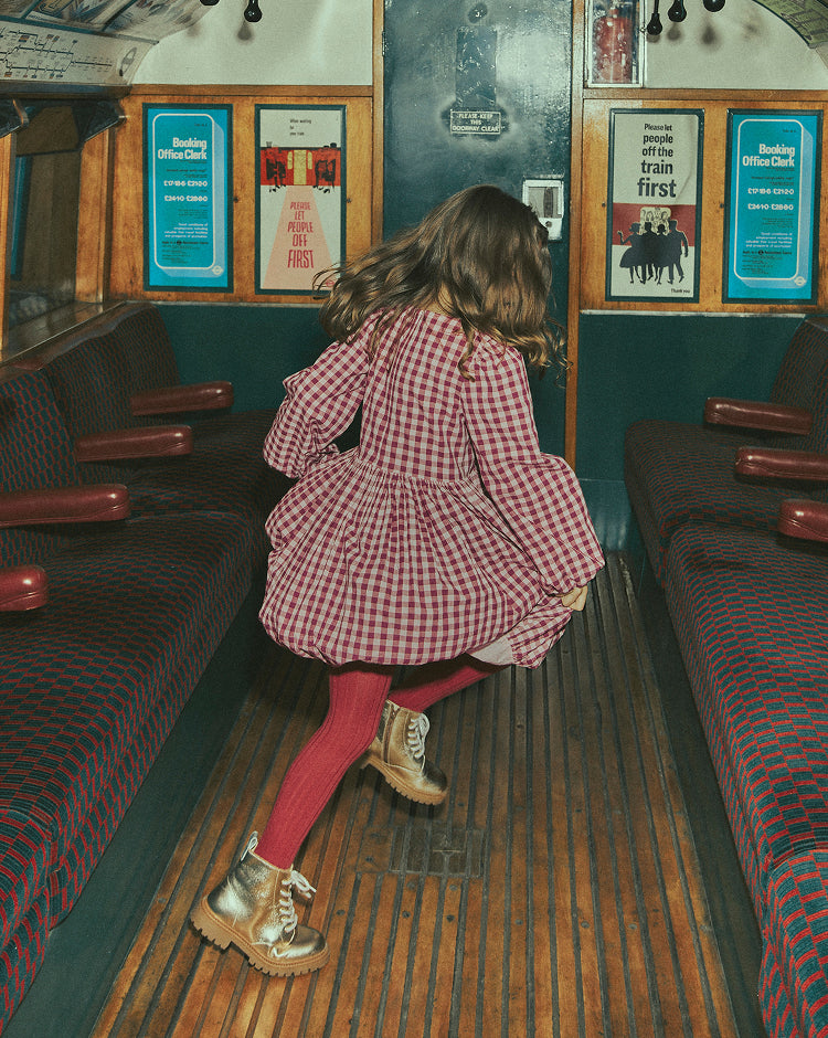 A young girl wearing a red and white gingham dress, pink tights, and gold boots stands in the aisle of a vintage train carriage, looking back over her shoulder. The train interior features wooden floors, red upholstered seats, and vintage posters on the walls.