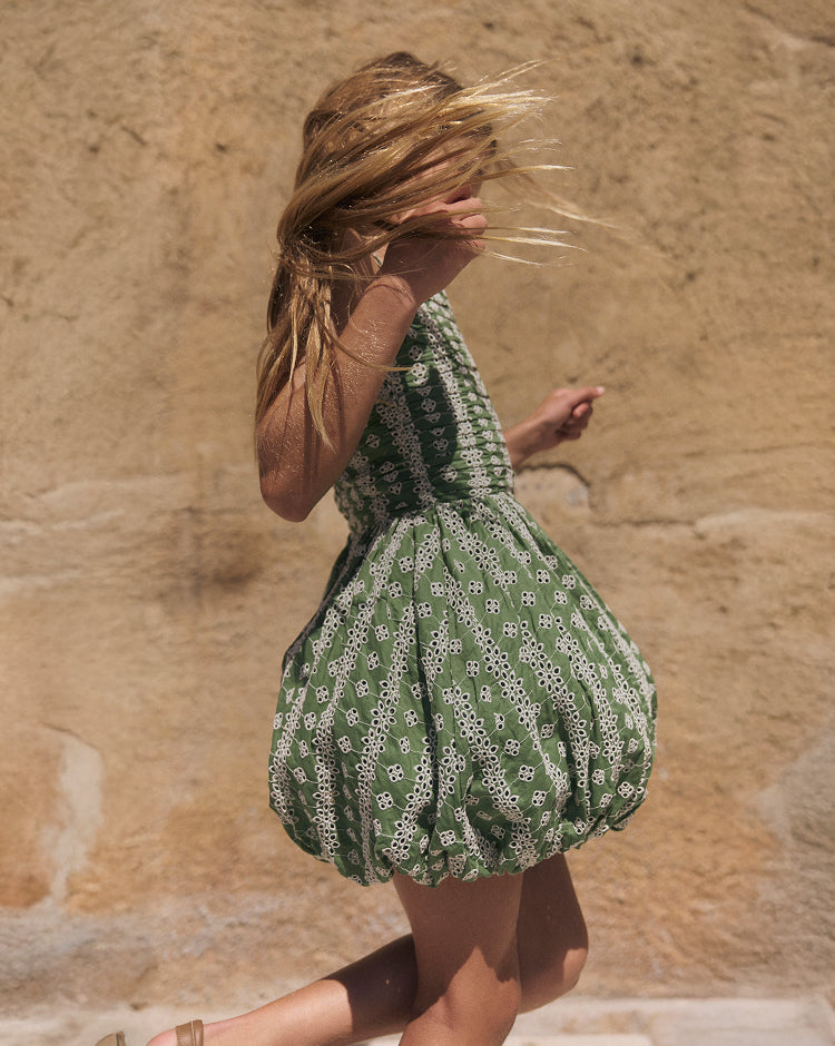 Woman in a green dress with white patterns standing against a beige stone wall.