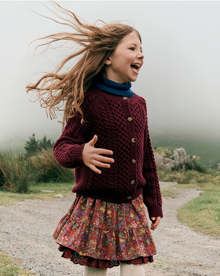 Young girl in a maroon sweater and floral skirt standing outdoors with windblown hair.