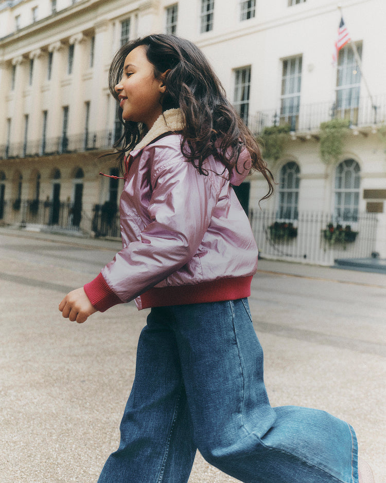 A young girl walking outdoors on a city street wearing a shiny light pink puffer jacket with red ribbed trim and blue wide-leg jeans.