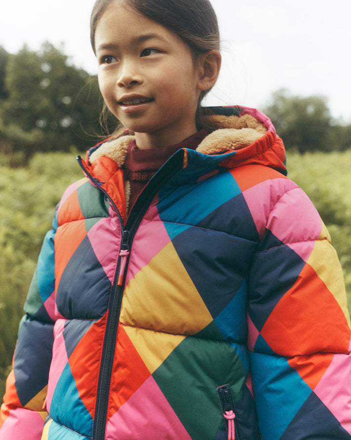 Close-up of a child outdoors wearing a colourful geometric puffer jacket in pink, orange, blue, green and yellow. The hood is lined with soft cream fleece, with a blurred green countryside landscape in the background.