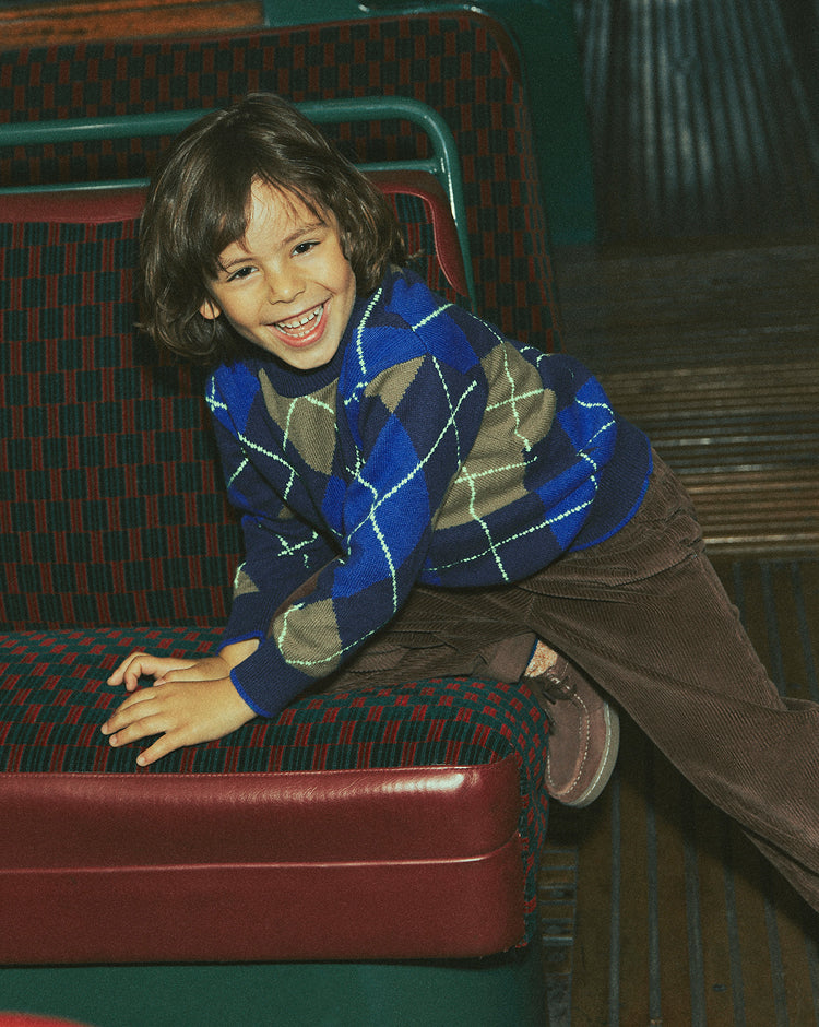 A young boy smiling while wearing a blue, brown, and beige argyle sweater and brown corduroy pants, sitting on a red and green patterned leather seat.