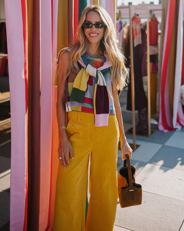 Women wearing sunglasses, mustard yellow trousers and a colourful striped top with a coordinating knit draped over the shoulders, standing among vibrant fabric panels.