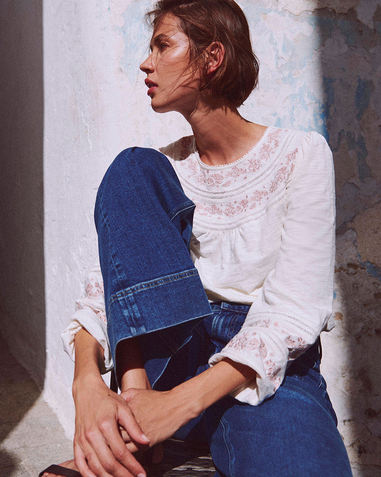 Woman wearing a white lace top and blue jeans sitting against a textured wall.