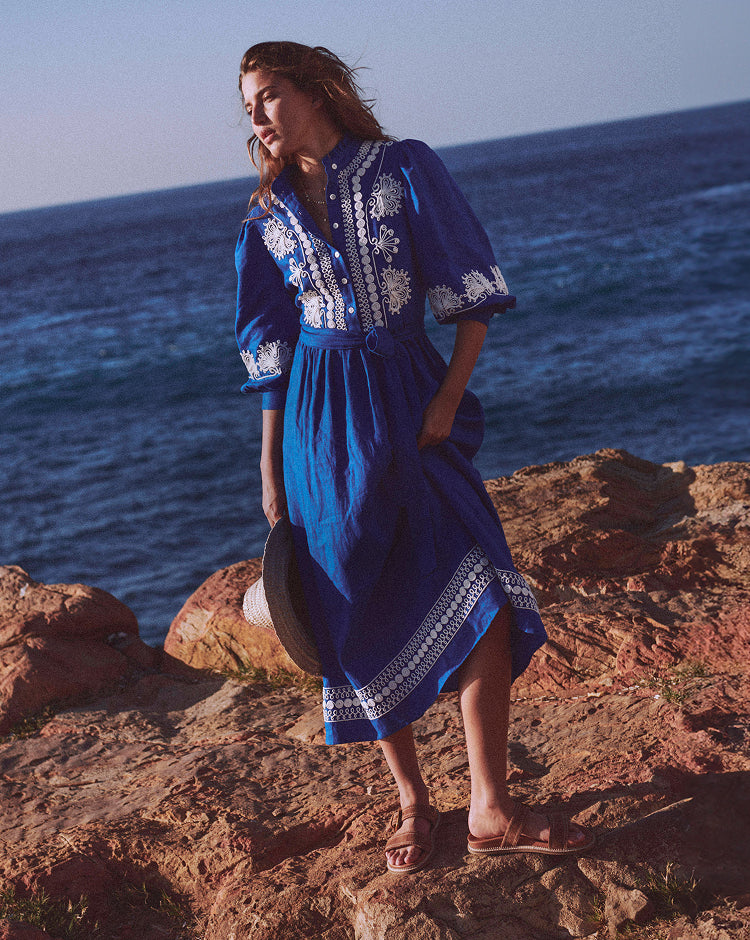 Woman in a blue dress with white embroidery standing on a rocky cliff overlooking the ocean.
