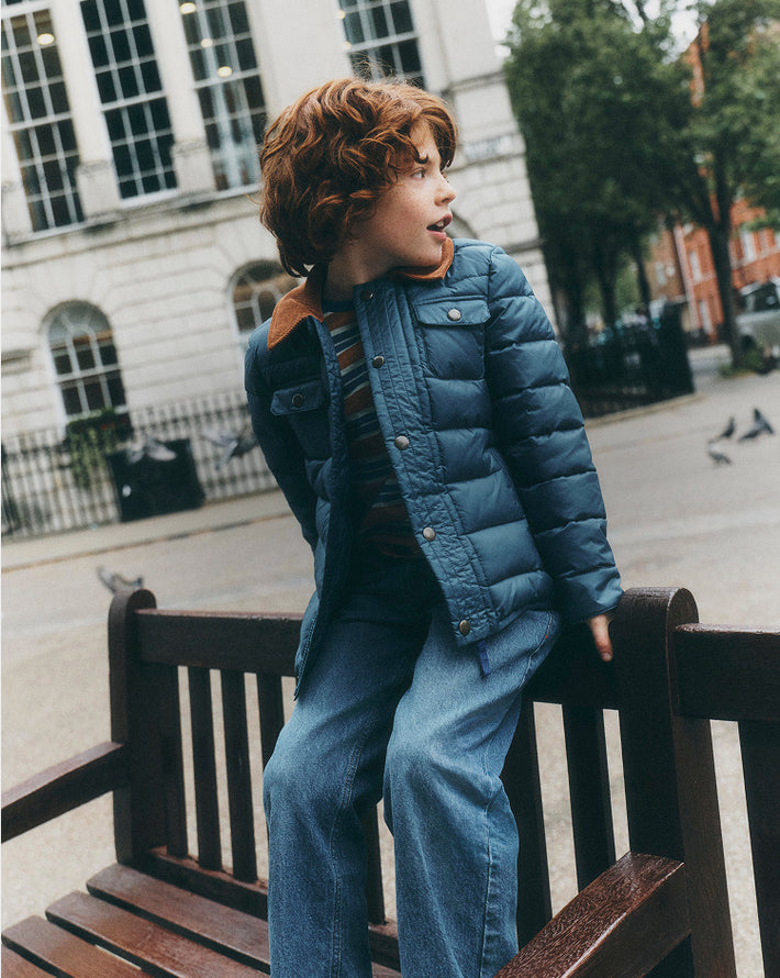 A young boy with curly red hair wearing a blue puffer jacket and matching blue jeans, sitting on a wooden bench in an outdoor urban setting.