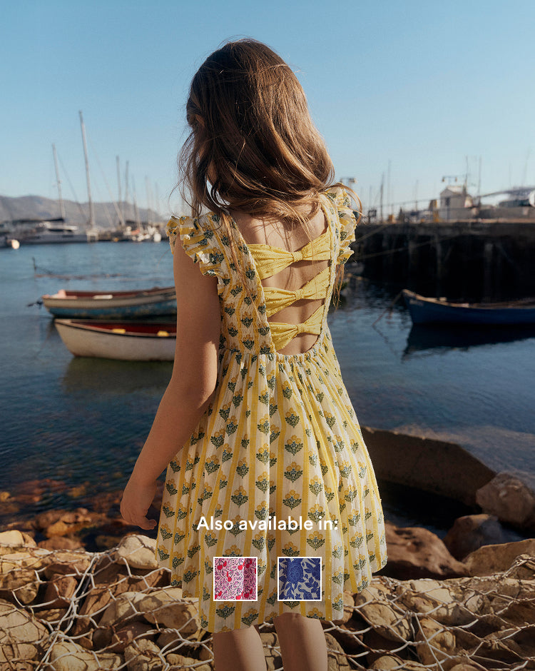 Girl in a yellow dress standing by a harbor with boats and netting on the ground.