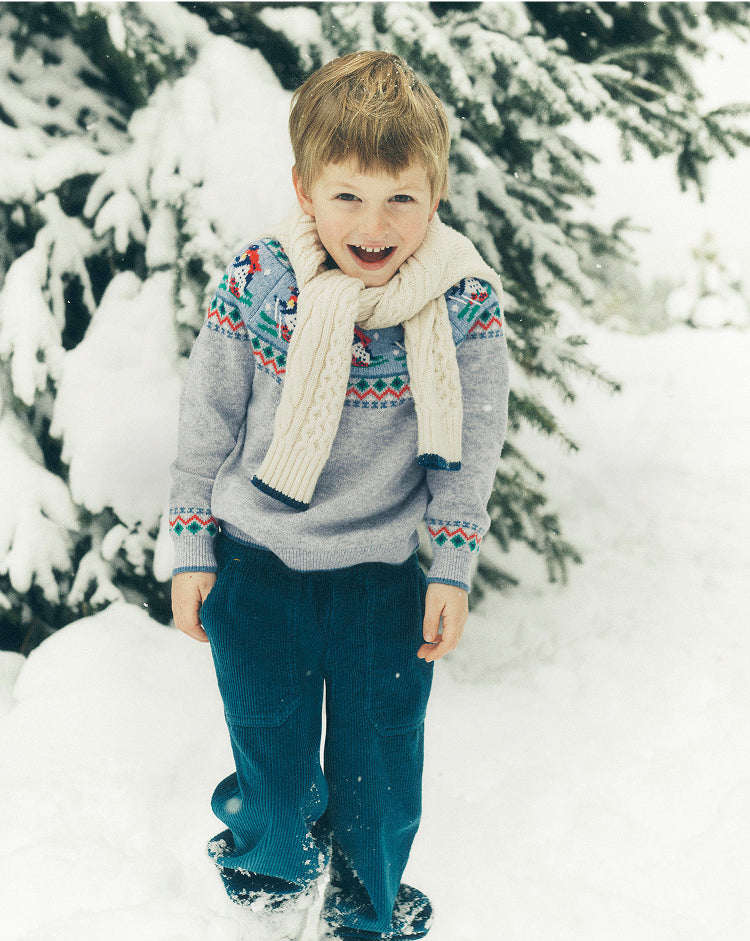 Child wearing a patterned sweater and scarf standing in the snow with trees in the background