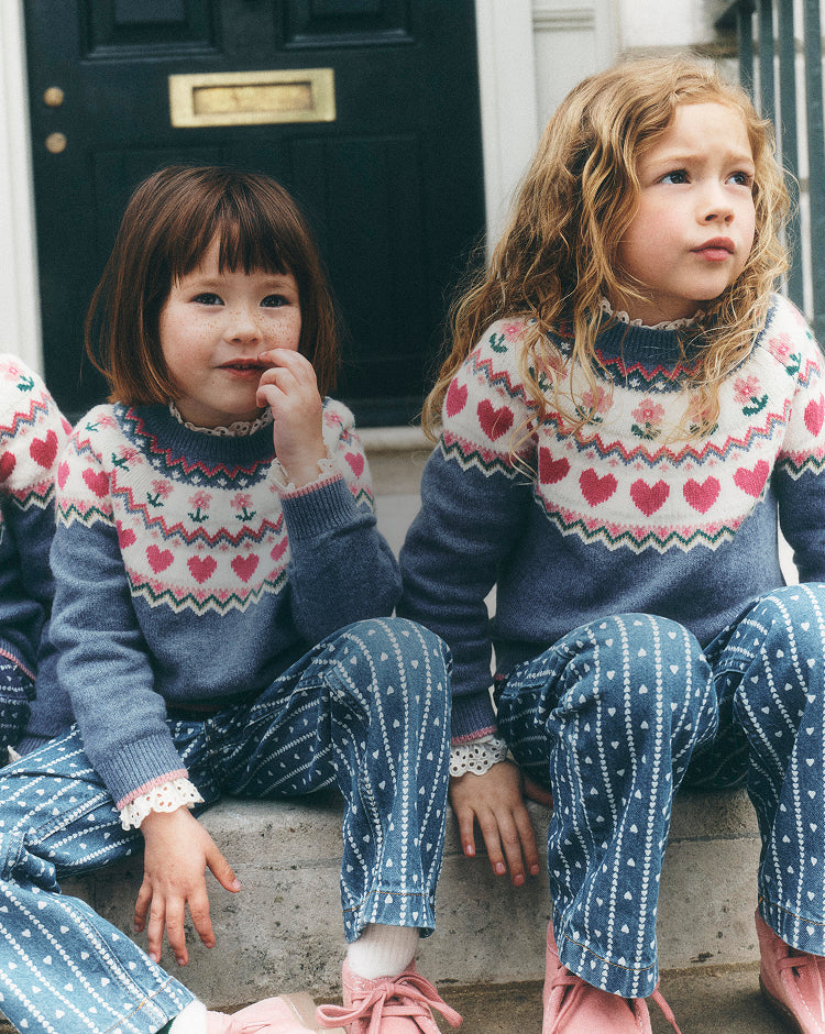 Two young girls sitting on stone steps in front of a dark door, wearing matching knit sweaters and patterned pants in a coordinated outfit.