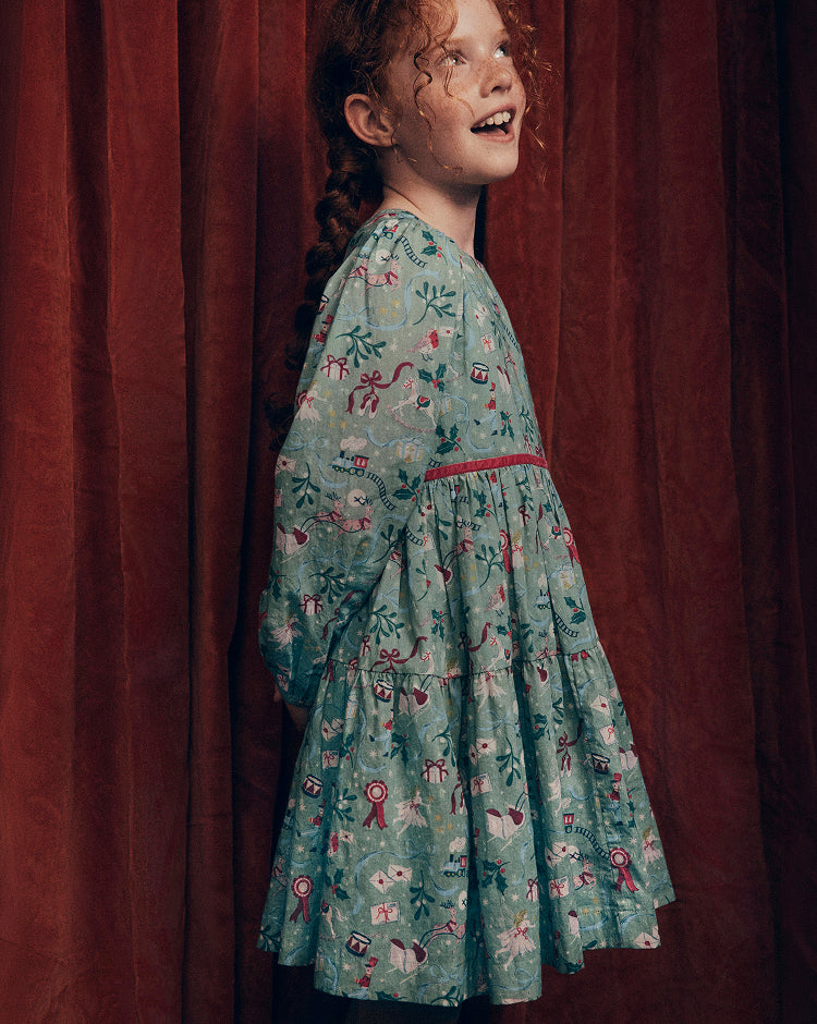 Child wearing a green long-sleeve dress with a festive illustrated print, gathered waist and tiered skirt