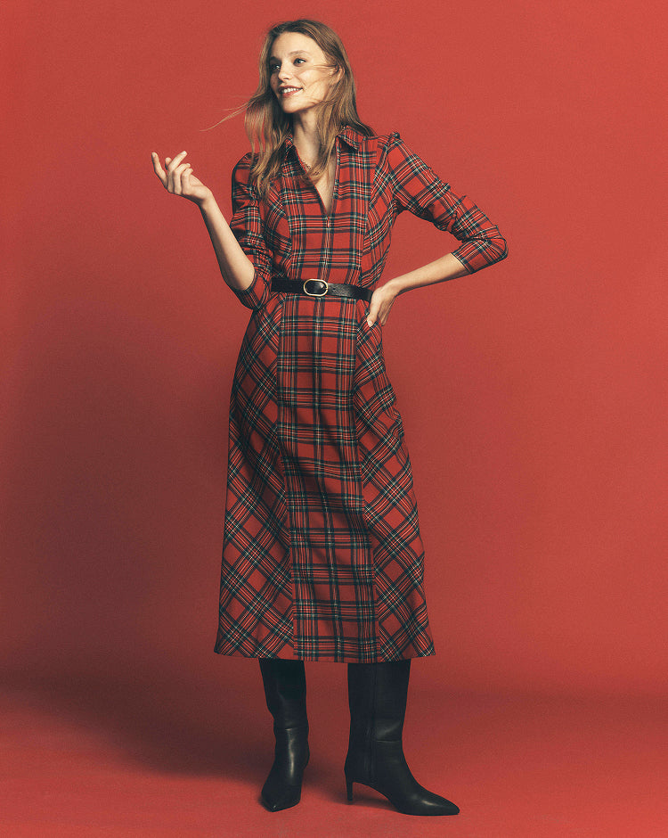 A woman modeling a red and black plaid midi shirt dress with a black belt, paired with black ankle boots, set against a solid red studio background.