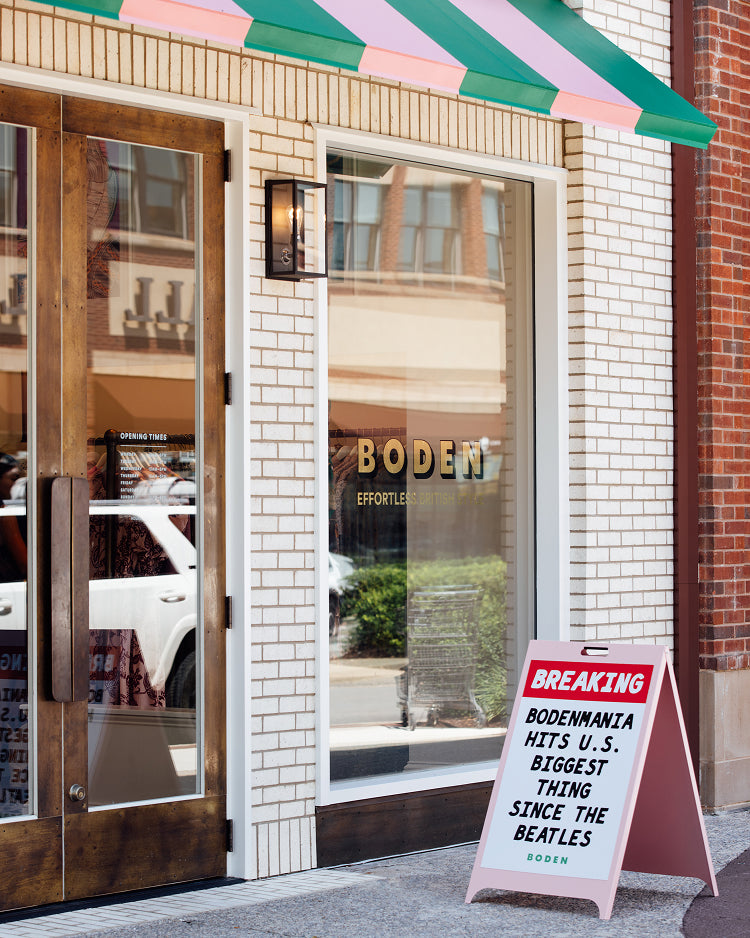 Boden Womanswear storefront entrance with a pink, green, and white striped awning, white brick facade, glass doors, and a sidewalk A-frame sign reading 'BREAKING BODENWANTIA HITS U.S.